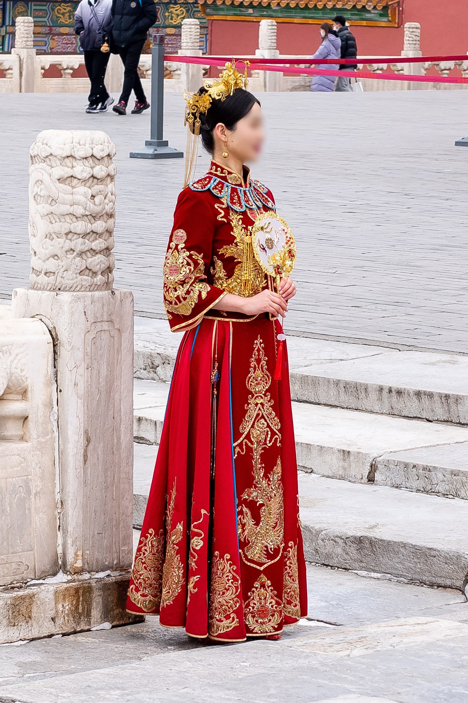 Jeune mariee en xiuhefu rouge brode de phenix d'or, coiffee d'ornements dores et tenant un eventail rond, posant sur le parvis dalle de la Cite interdite a Pekin