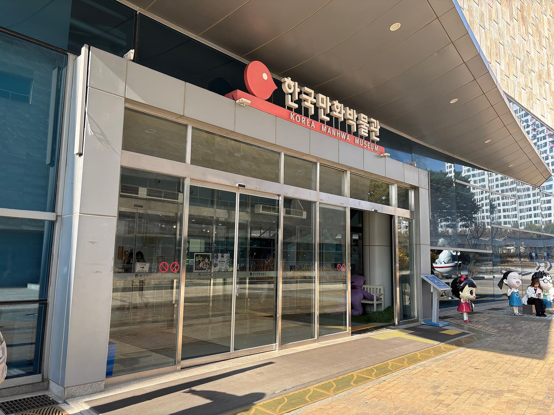 Entree vitree du Musee coreen du manhwa a Bucheon, enseigne rouge portant l'inscription en hangeul 한국만화박물관 et sa traduction Korea Manhwa Museum, avec des statues de personnages de bande dessinee coreenne postees sur le parvis