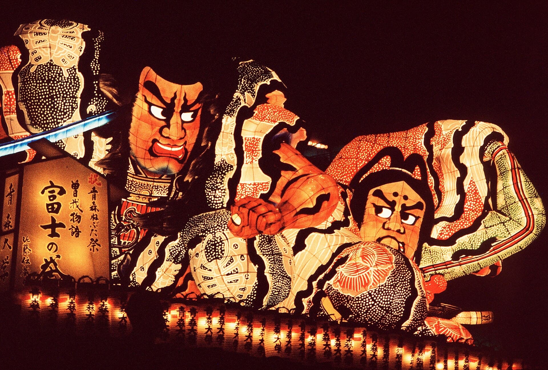 Char-lanterne du Nebuta Matsuri d'Aomori en défilé nocturne : deux guerriers de papier washi illuminés de l'intérieur, avec un panneau portant les kanji du festival.