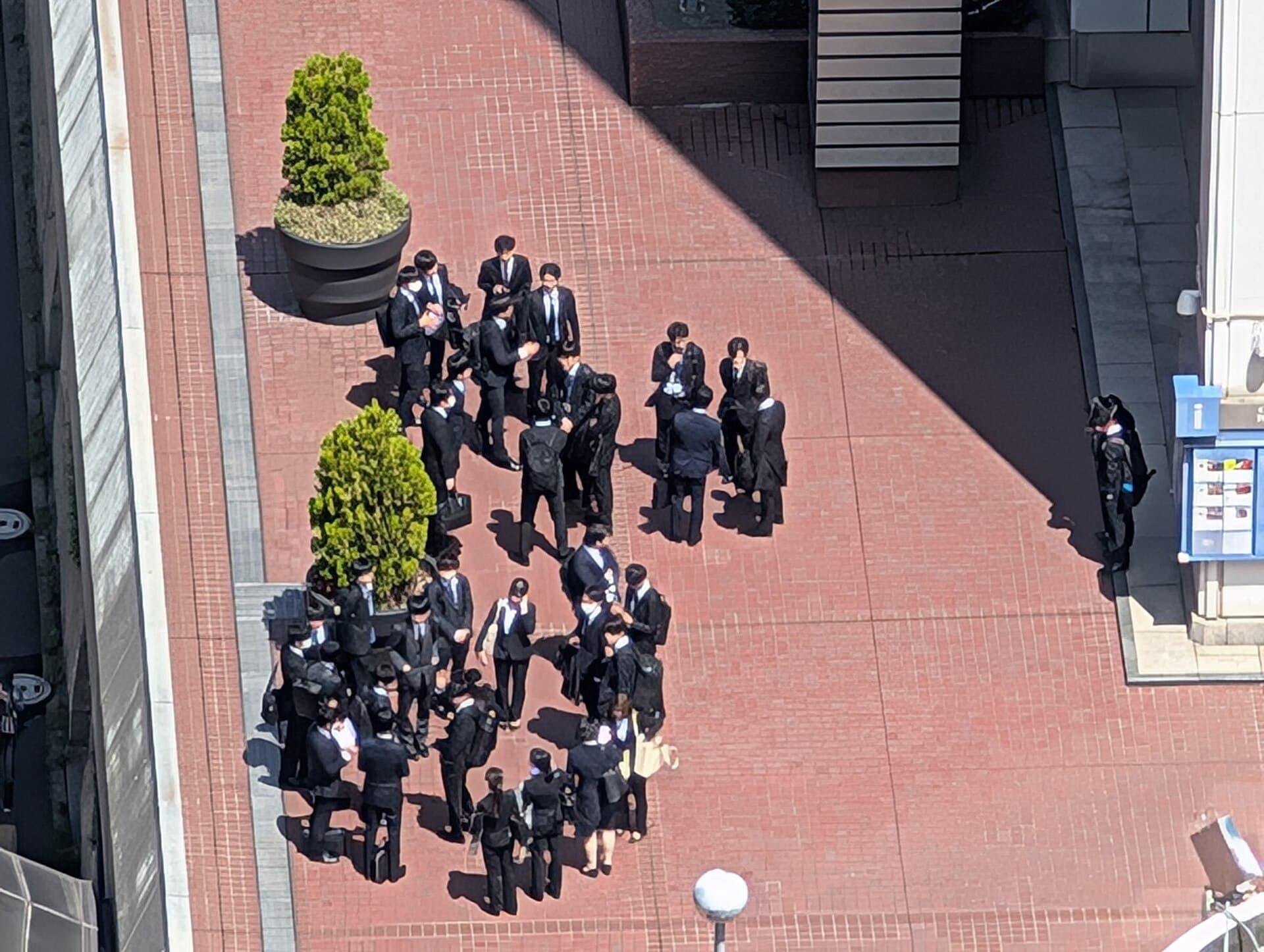 Groupe de jeunes salariés japonais en costume (nouvelles recrues, shinnyū shain) rassemblés devant une tour de bureaux à Tōkyō.