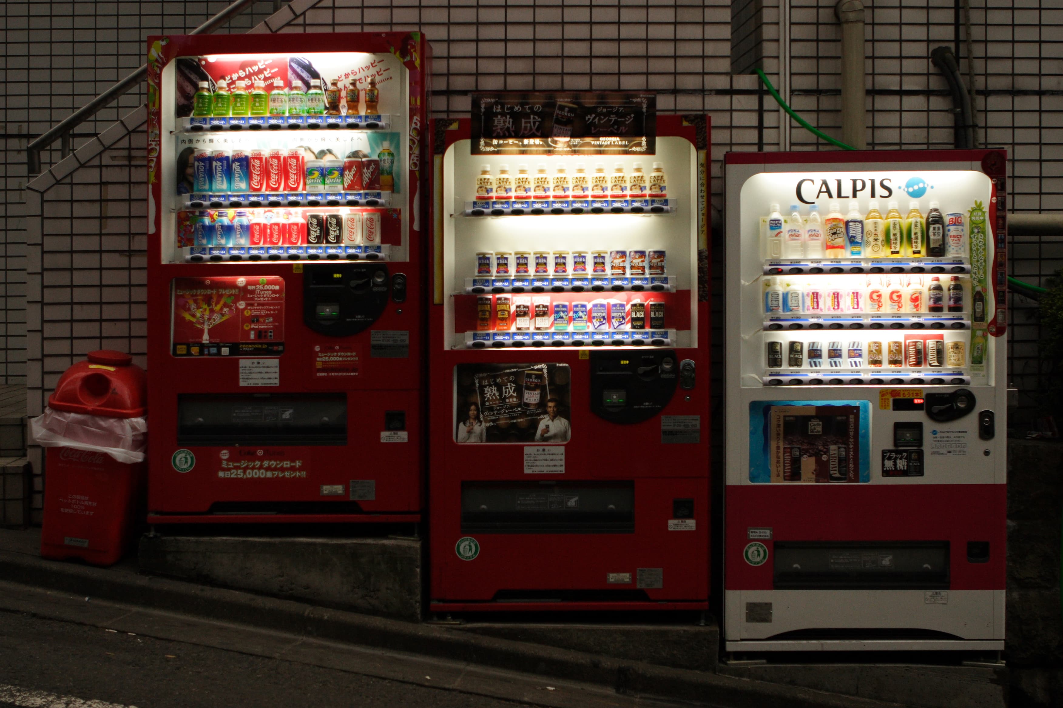 Trois distributeurs automatiques de boissons alignés le long d'une rue japonaise, éclairés la nuit.