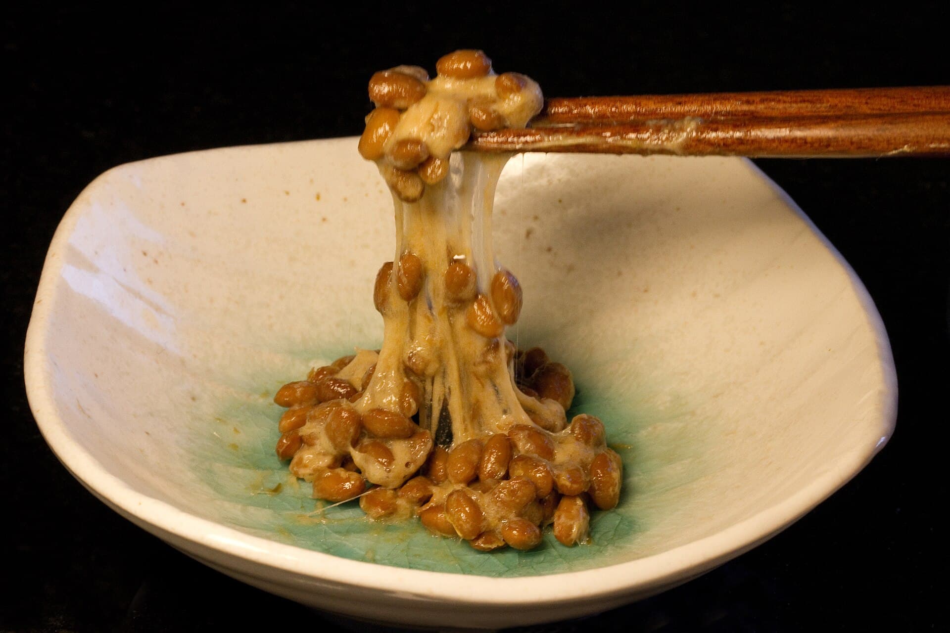 Gros plan sur un bol japonais creme contenant du natto : une paire de baguettes souleve des graines de soja fermentees reliees au bol par de longs filaments blancs et elastiques caracteristiques de la fermentation par Bacillus subtilis