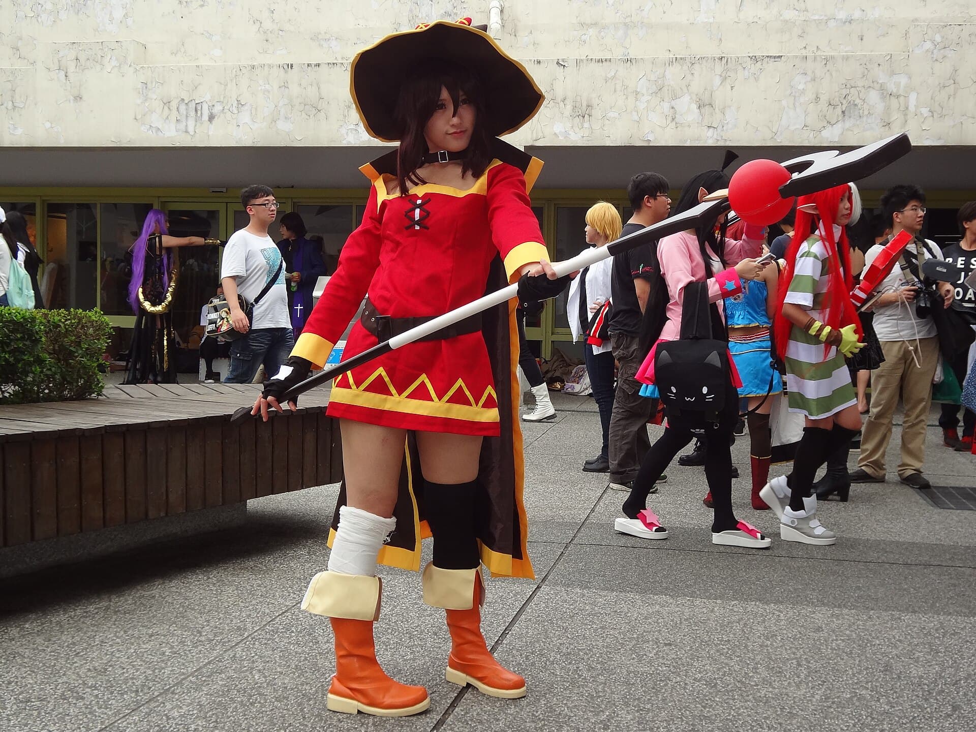 Cosplayeuse incarnant Megumin, la magicienne de la serie isekai KonoSuba : robe rouge a lisere jaune, grand chapeau noir a large bord, cape sombre et long baton tenu a deux mains, photographiee en exterieur lors d'une convention