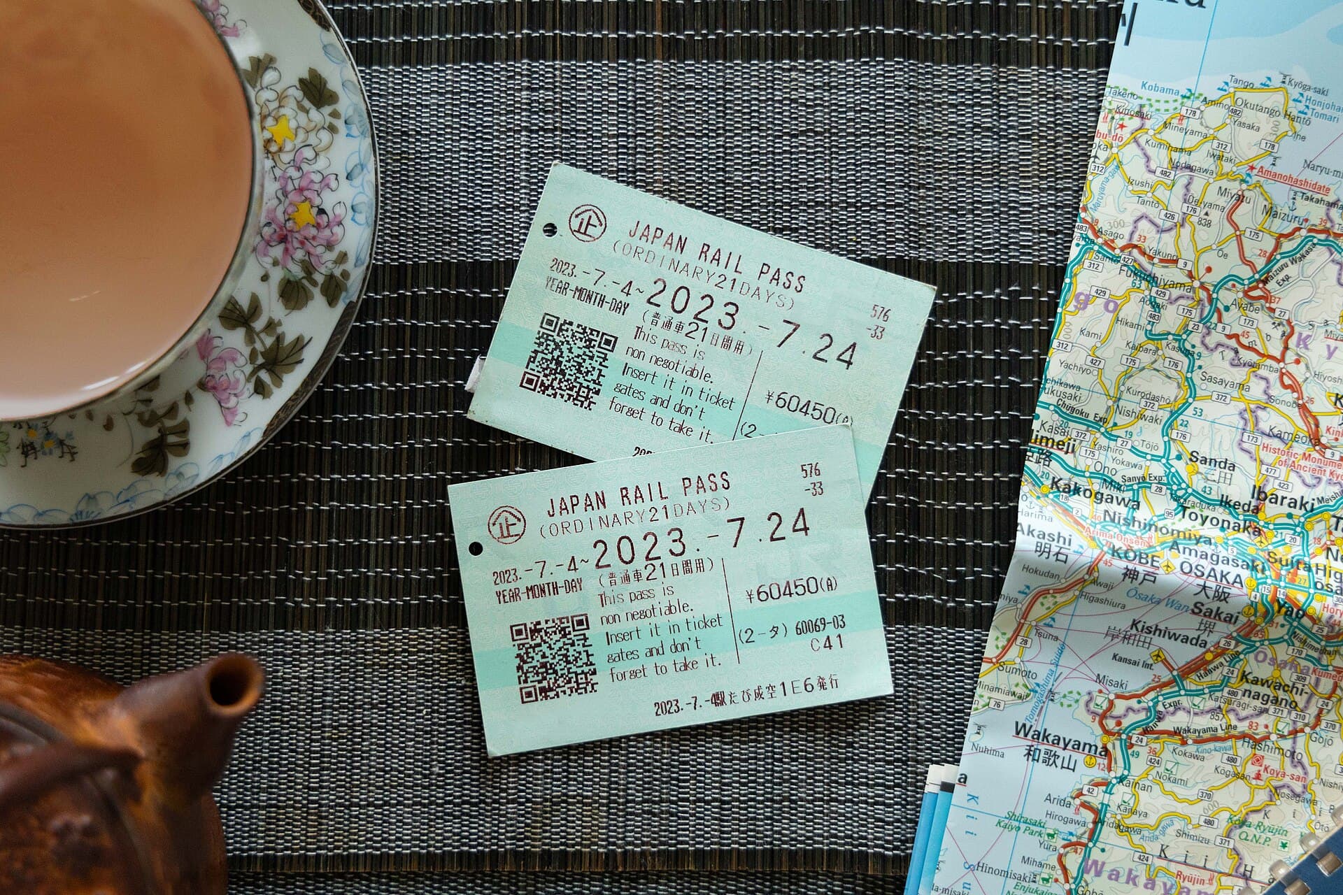 Deux Japan Rail Pass ordinaires de 21 jours poses sur un set de table sombre, tampons de validite de juillet 2023 et prix de 60 450 yens imprimes en rouge, a cote d'une carte routiere du Kansai depliee et d'une tasse de the