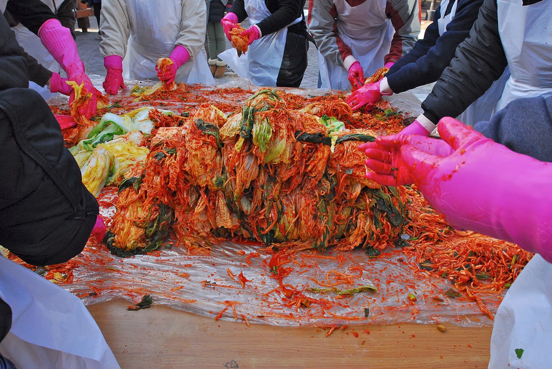 Scène de kimjang en Corée : plusieurs personnes agenouillées préparent ensemble du kimchi d'hiver, enduisant des choux napa de pâte de piment rouge dans de grandes bassines lors d'une préparation collective au village de Gaemi.