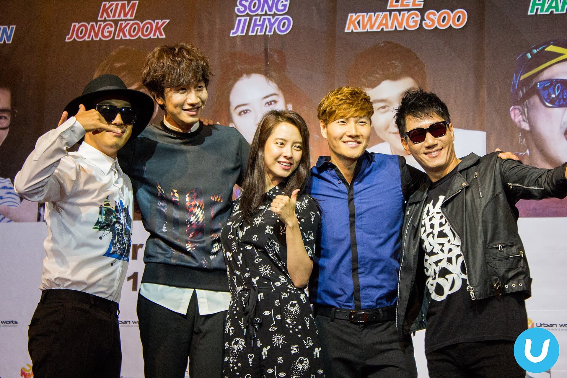 Cinq membres de l'emission Running Man poses bras dessus bras dessous devant le decor d'une rencontre avec les fans en 2014, les prenoms des participants imprimes en grandes lettres colorees derriere eux